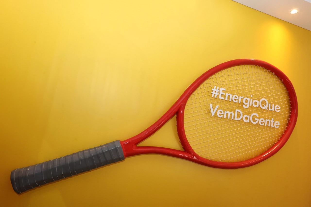 Raquete de tênis vermelha instalada como objeto decorativo em uma parede amarela, com a hashtag ‘#EnergiaQueVemDaGente’ escrita sobre as cordas.