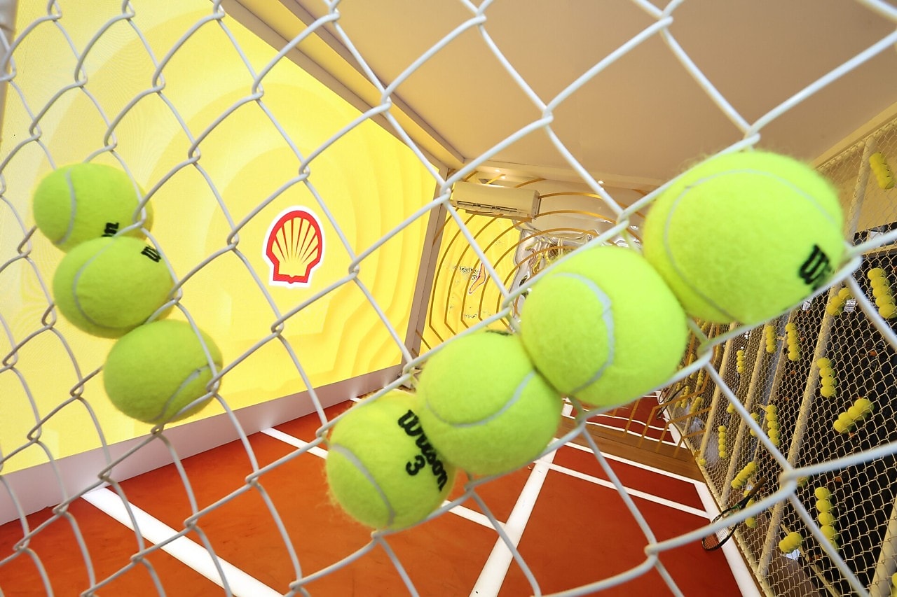 Bolas de tênis presas a uma estrutura metálica, com o logotipo da Shell ao fundo em um ambiente decorado com temática esportiva.