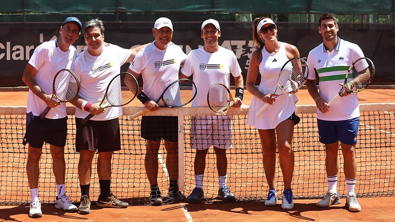 Grupo de adultos vestindo roupas esportivas posa em uma quadra de tênis de saibro, segurando raquetes e sorrindo para a câmera, com a rede ao centro da imagem 