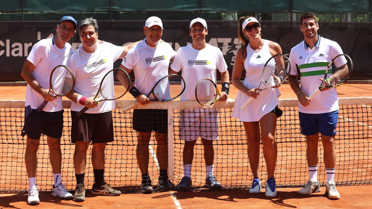 Grupo de adultos vestindo roupas esportivas posa em uma quadra de tênis de saibro, segurando raquetes e sorrindo para a câmera, com a rede ao centro da imagem 