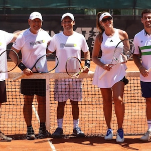 Grupo de adultos vestindo roupas esportivas posa em uma quadra de tênis de saibro, segurando raquetes e sorrindo para a câmera, com a rede ao centro da imagem 