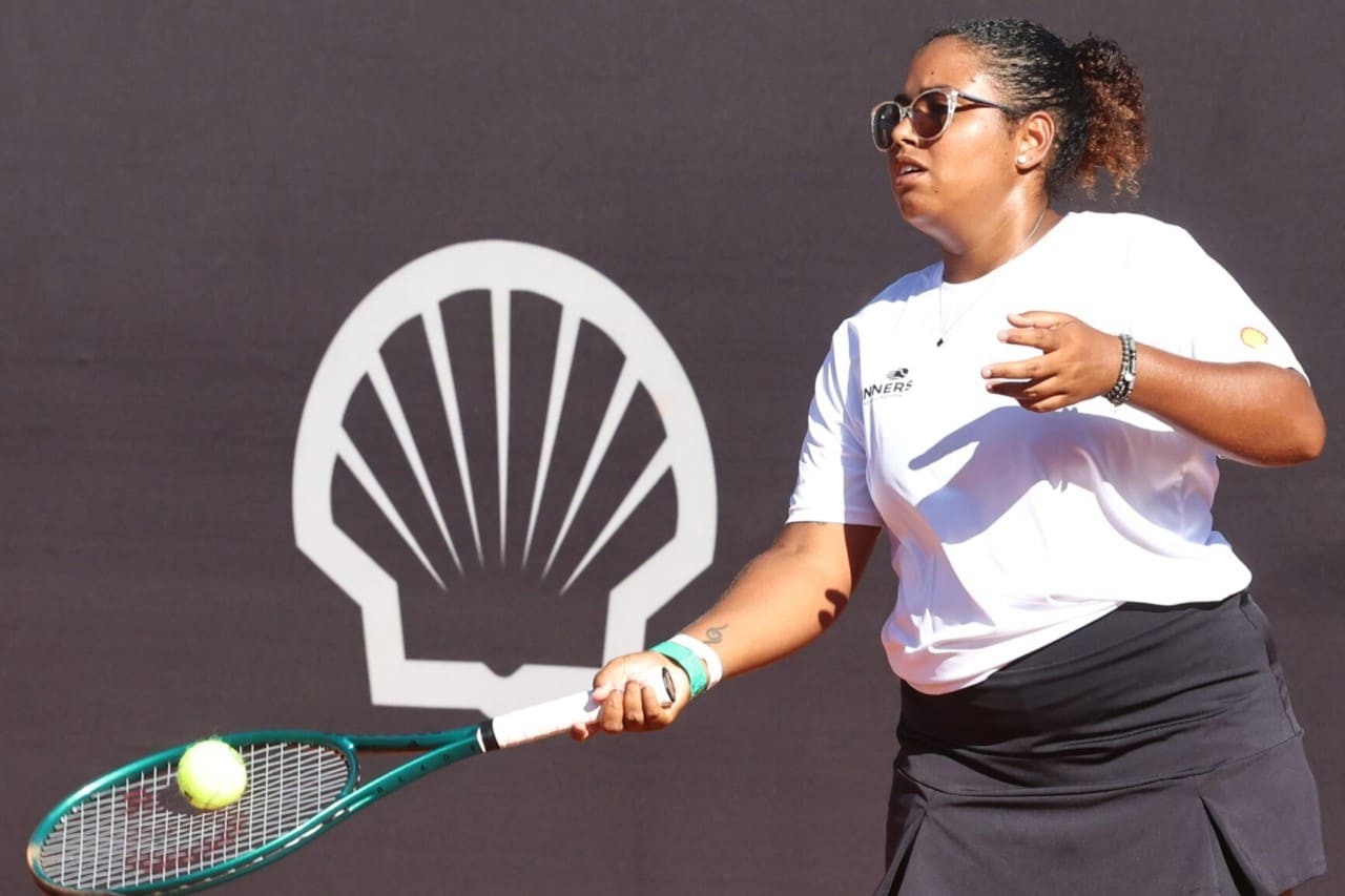 Mulher jogando tênis em quadra de saibro, segurando a raquete enquanto acompanha a bola, com o logotipo da Shell ao fundo.