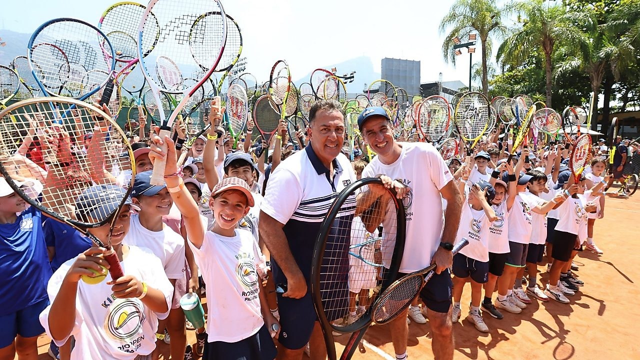 Grande grupo de crianças e adultos em uma quadra de tênis levanta raquetes para o alto em gesto de celebração, durante um evento esportivo ao ar livre.