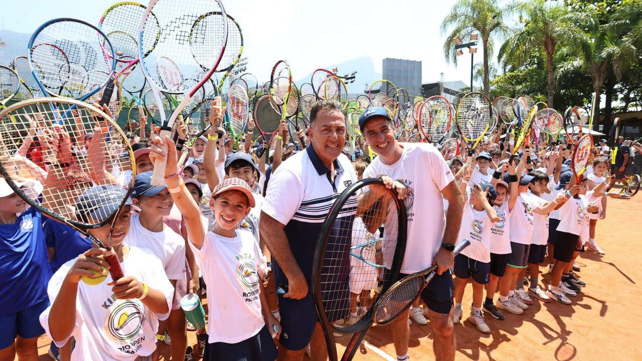 Grande grupo de crianças e adultos em uma quadra de tênis levanta raquetes para o alto em gesto de celebração, durante um evento esportivo ao ar livre.