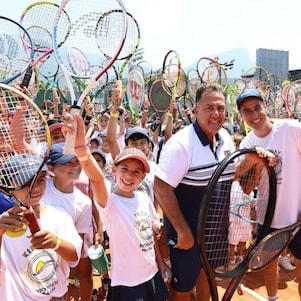 Grande grupo de crianças e adultos em uma quadra de tênis levanta raquetes para o alto em gesto de celebração, durante um evento esportivo ao ar livre.