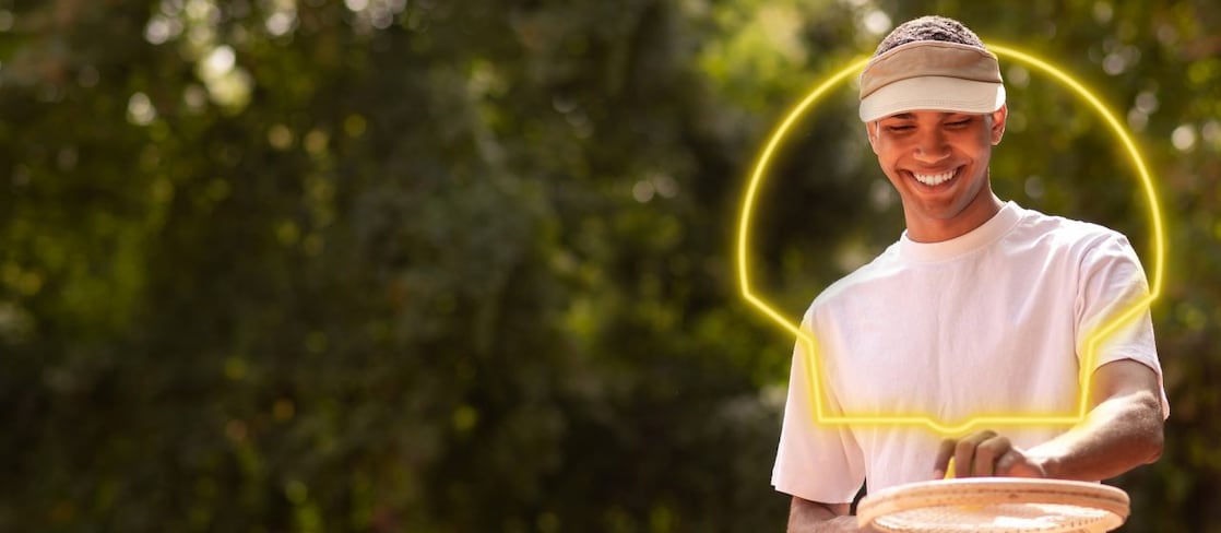 na imagem, um rapaz negro, jovem e de camiseta branca está sorrindo com sua raquete e bolinha de tênis.