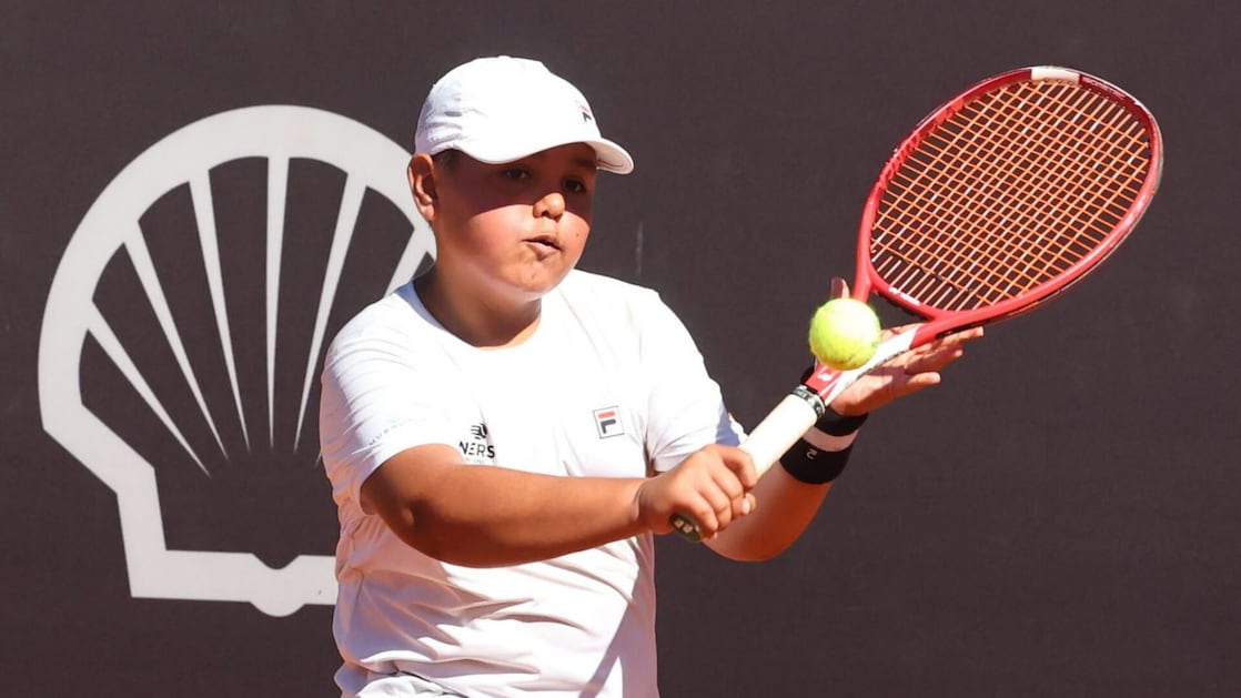 Criança jogando tênis em uma quadra, segurando uma raquete vermelha e rebatendo uma bola verde. A criança usa camiseta branca, boné branco e pulseira esportiva, com um fundo escuro ao fundo.