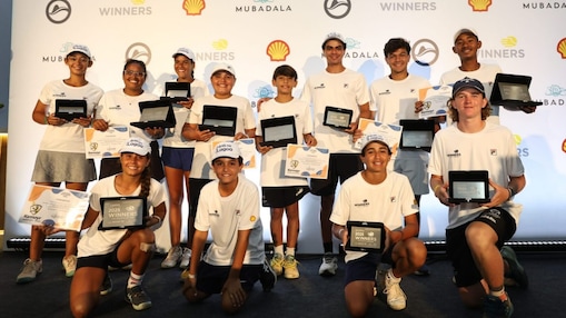 Grupo de crianças e adolescentes vestindo camisetas brancas posa para foto em evento esportivo, segurando troféus e certificados. Ao fundo, há um painel com logos de patrocinadores, como Shell e Mubadala, indicando uma cerimônia de premiação.