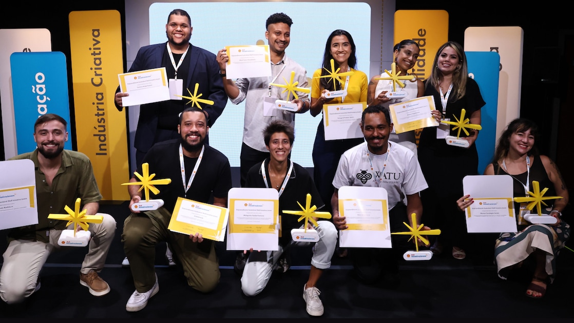 Um grupo de onze pessoas posa sorrindo em um palco durante uma cerimônia de premiação. Todas seguram certificados e troféus amarelos em forma de estrela, indicando que foram reconhecidas ou vencedoras. Elas estão organizadas em duas fileiras – cinco pessoas em pé atrás e seis agachadas ou sentadas à frente. Ao fundo, há painéis verticais coloridos com palavras como “Indústria Criativa”, “Inovação” e “Criatividade”. O clima é celebrativo, com todos demonstrando orgulho e alegria pela conquista.