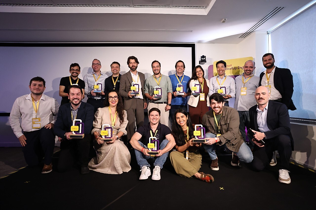 Grupo de participantes do programa Shell StartUp Energy posa sorridente em um auditório. As pessoas seguram troféus coloridos e usam crachás amarelos do evento. Ao fundo, há uma tela de projeção e um banner do programa, em um ambiente iluminado e profissional.