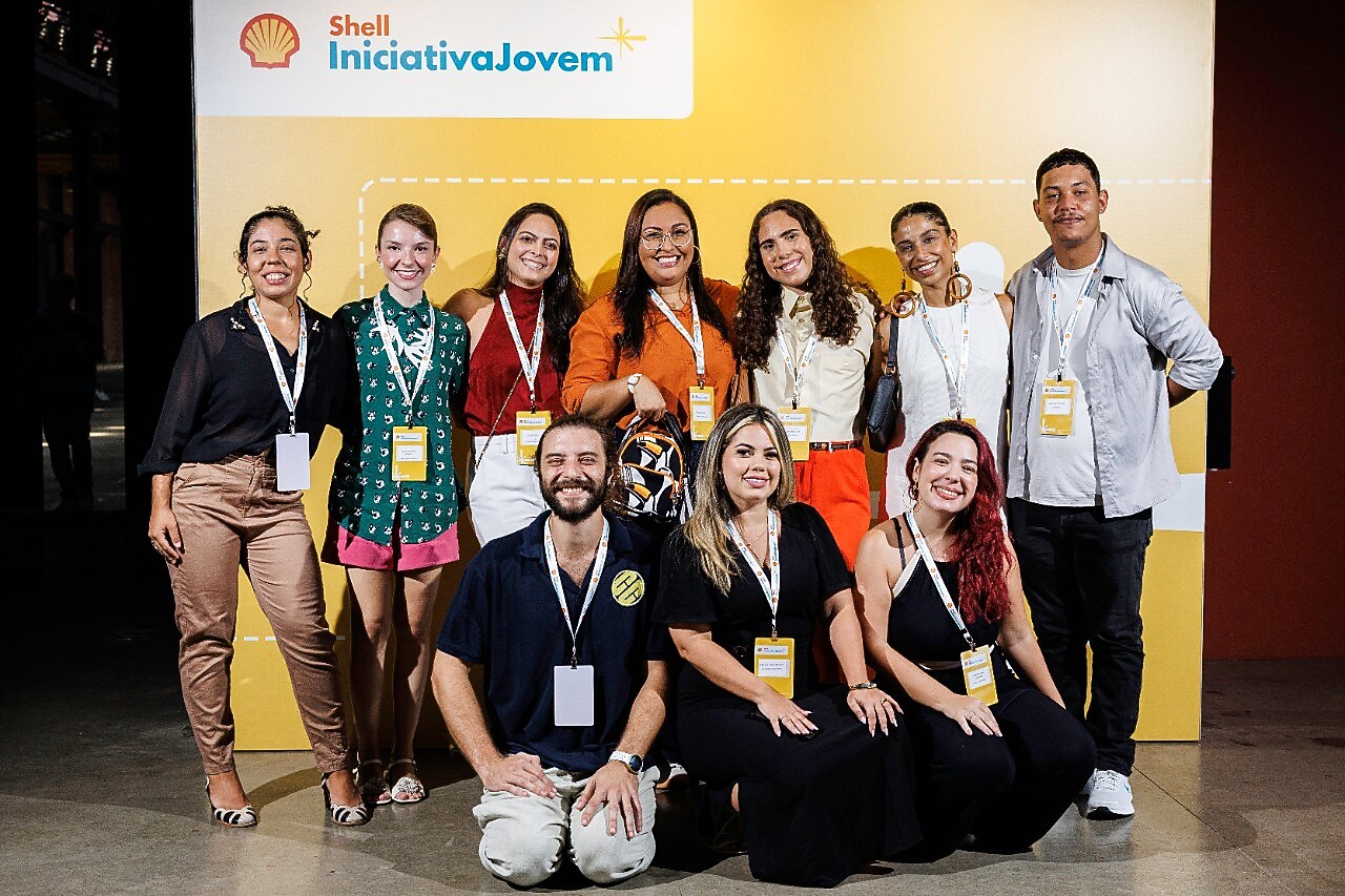 Grupo de participantes do Shell Iniciativa Jovem posa sorridente em frente a um painel amarelo com o logotipo do programa. Nove pessoas estão de pé e três agachadas à frente, todas usando crachás de identificação e roupas casuais coloridas, em um ambiente iluminado e descontraído.