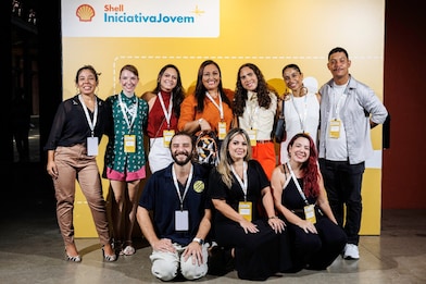 Grupo de participantes do Shell Iniciativa Jovem posa sorridente em frente a um painel amarelo com o logotipo do programa. Nove pessoas estão de pé e três agachadas à frente, todas usando crachás de identificação e roupas casuais coloridas, em um ambiente iluminado e descontraído.