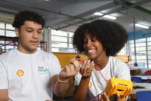 Dois jovens usam camisetas brancas do programa Shell NXplorers e trabalham juntos em uma atividade prática de ciências. Eles sorriem enquanto manuseiam cabos e um multímetro em uma sala de aula colorida e iluminada.