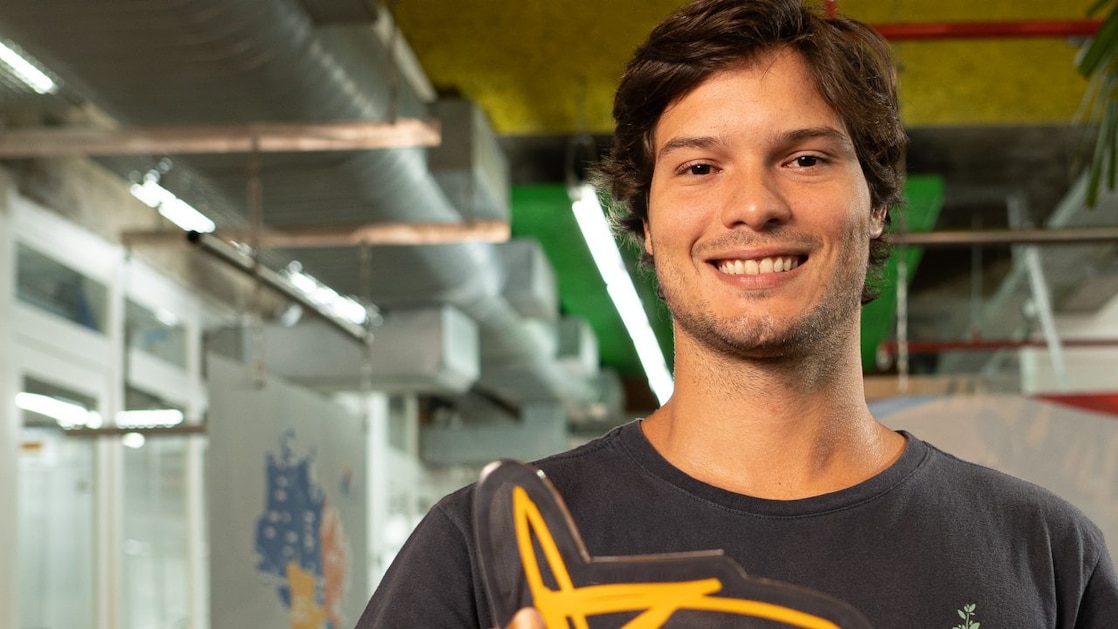 Um homem de blusa preta, segurando uma placa amarela e  sorrindo.