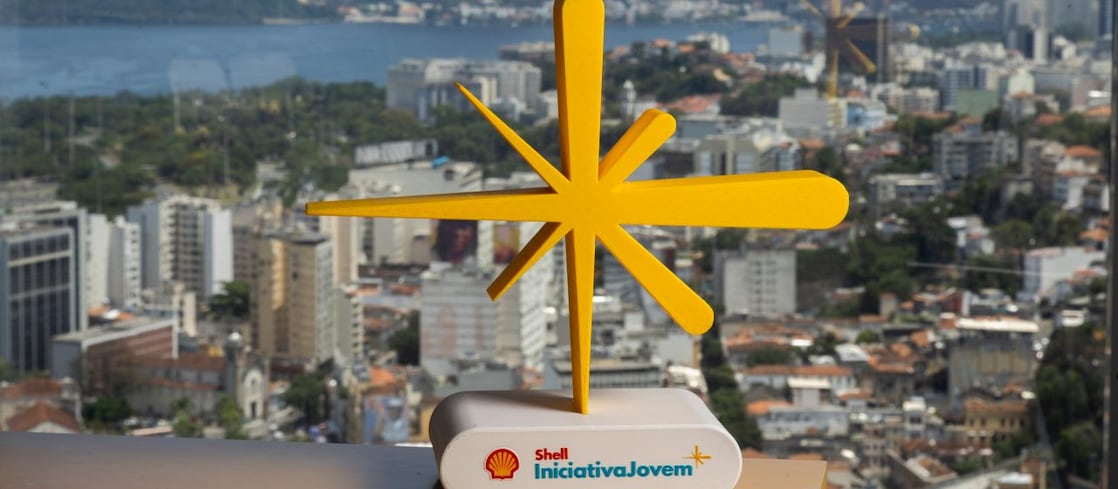 Escultura amarela em forma de estrela sobre base branca com o logotipo Shell Iniciativa Jovem, posicionada diante de uma vista urbana do Rio de Janeiro, com prédios, área verde, o mar e o Pão de Açúcar ao fundo.