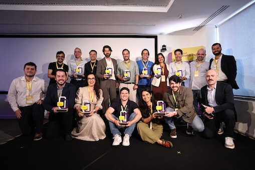 Grupo de participantes do programa Shell StartUp Energy posa sorridente em um auditório. As pessoas seguram troféus coloridos e usam crachás amarelos do evento. Ao fundo, há uma tela de projeção e um banner do programa, em um ambiente iluminado e profissional.