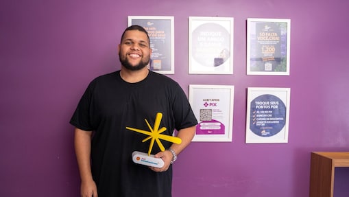 Pessoa sorrindo em ambiente interno, em frente a uma parede roxa com quadros informativos ao fundo, segura um troféu amarelo do Shell Iniciativa Jovem sobre base branca, usando camiseta preta e posando de frente para a câmera.