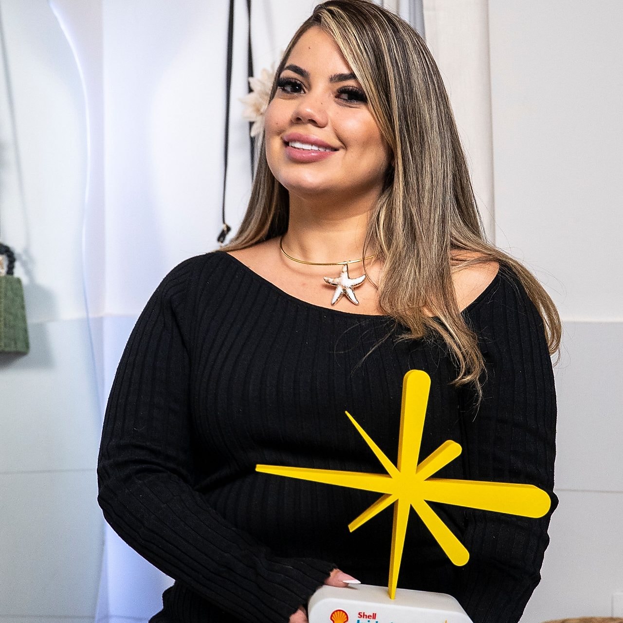 Mulher sentada, sorrindo para a câmera, usando vestido preto de manga longa e colar em formato de estrela-do-mar, segura um troféu branco com o logo do Shell Iniciativa Jovem e uma escultura amarela em formato abstrato; ao fundo, ambiente interno claro com objetos decorativos.