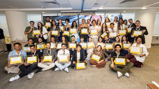 Grupo diverso de participantes posa em ambiente corporativo durante cerimônia do Shell Iniciativa Jovem, segurando certificados e sorrindo para a câmera, com painel do programa ao fundo e pessoas sentadas e em pé organizadas em fileiras.