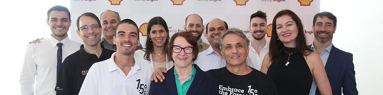 Grupo de 13 pessoas posando sorridentes em frente a um painel com o logo da Shell e a marca Shell StartUp Engine. A foto mostra diversidade de gênero e estilo, com pessoas vestindo roupas sociais. O ambiente é iluminado e transmite clima de celebração e colaboração