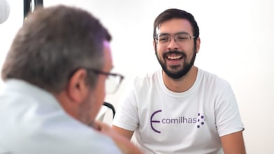 Homem jovem com barba e óculos, vestindo camiseta branca com o logo da Comilhas, sorri durante uma conversa com outro homem de cabelos grisalhos em primeiro plano, em um ambiente iluminado e descontraído