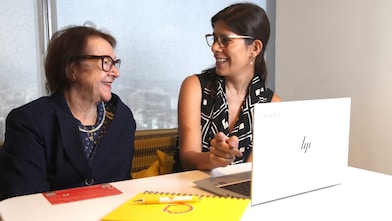 Mulher jovem e mulher idosa conversam sorridentes em uma mesa de escritório. A jovem segura uma caneta e está à frente de um notebook HP aberto. Sobre a mesa há um caderno amarelo com um símbolo de lâmpada e um folheto vermelho. Ambas usam óculos e parecem estar em um ambiente corporativo iluminado naturalmente.