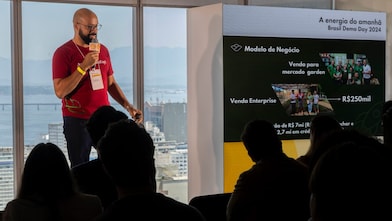 Homem negro com barba e óculos apresenta um pitch no evento "Brasil Demo Day 2024" da Shell, segurando um microfone com o logotipo da empresa. Ele veste uma camiseta vermelha com o texto “A energia do amanhã”. Ao fundo, uma janela com vista para a Baía de Guanabara e, ao lado, uma tela exibe informações sobre o modelo de negócio apresentado