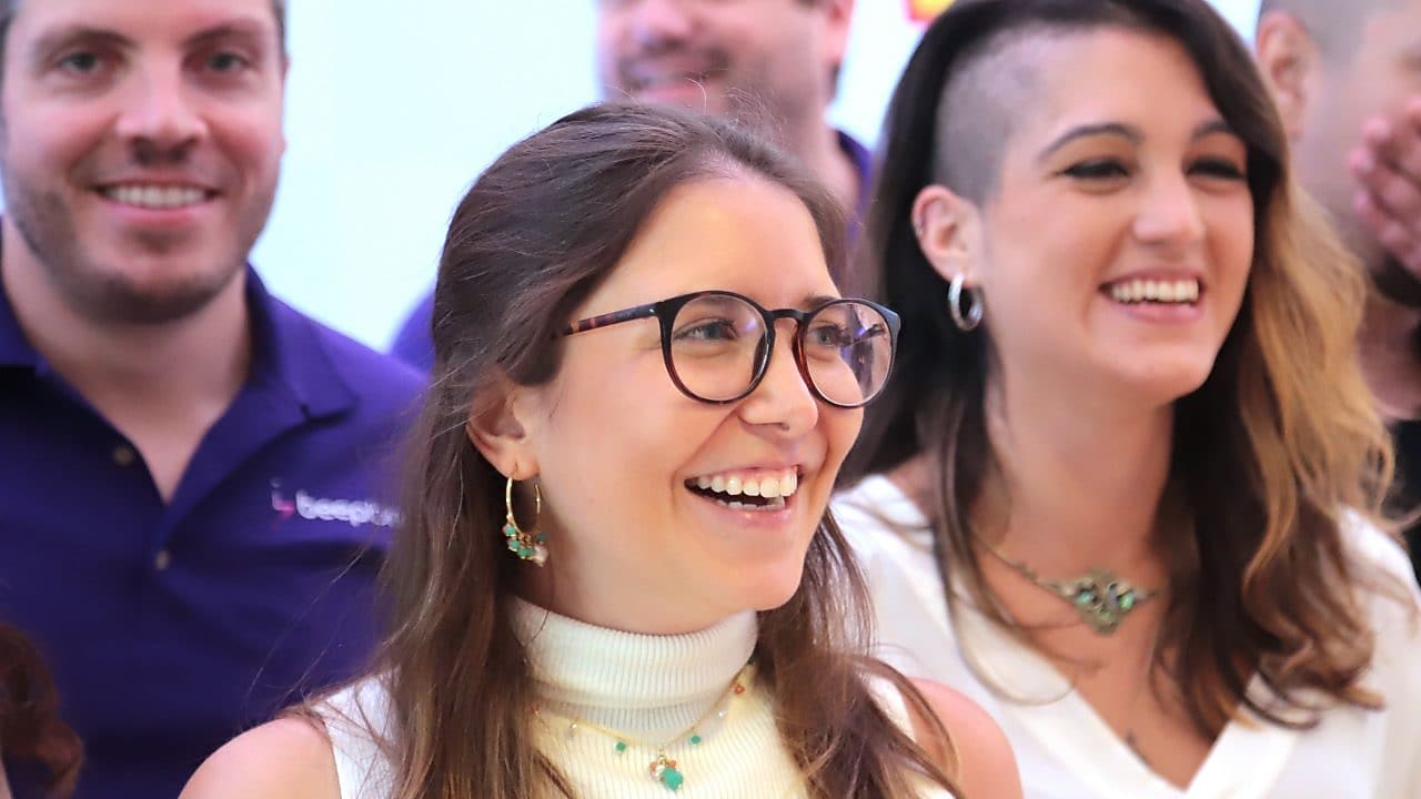 Grupo de pessoas sorrindo em um ambiente claro e descontraído. Em destaque, uma mulher com óculos, blusa branca sem mangas e joias turquesa, ao lado de outra mulher com cabelo raspado de um lado e blusa branca. Ao fundo, outras pessoas também sorriem, vestindo roupas casuais.
