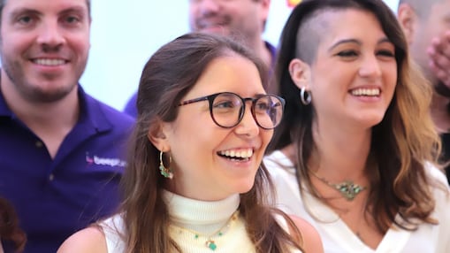 Grupo de pessoas sorrindo em um ambiente claro e descontraído. Em destaque, uma mulher com óculos, blusa branca sem mangas e joias turquesa, ao lado de outra mulher com cabelo raspado de um lado e blusa branca. Ao fundo, outras pessoas também sorriem, vestindo roupas casuais.