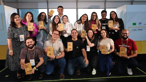 Grupo de pessoas sorridentes posando para foto segurando troféus de madeira, em um ambiente iluminado com fundo institucional. Todos usam crachás, indicando participação em um evento corporativo ou cerimônia de premiação.