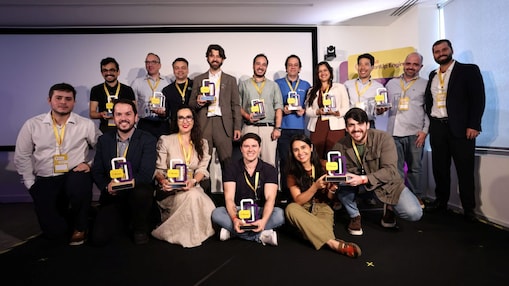 Grupo de cerca de vinte pessoas posando para foto em um ambiente corporativo, todas usando crachás amarelos. Eles estão organizados em duas fileiras, alguns em pé e outros agachados ou sentados à frente. Cada pessoa segura um troféu colorido, sugerindo uma premiação ou reconhecimento. Ao fundo, há uma tela branca de projeção e um painel com a identidade visual do evento.