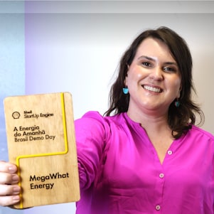 Mulher branca, de cabelos escuros, sorrindo enquanto segura um troféu de madeira.