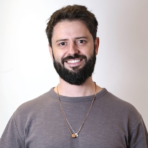 Homem, branco e de camisa cinza, sorrindo em um fundo branco.