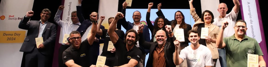 Grupo de cerca de 16 pessoas posa com expressão de comemoração no palco do evento “Shell StartUp Engine – Demo Day 2024”. Todos sorriem e levantam os punhos em sinal de vitória, segurando placas ou troféus. Alguns estão agachados na frente, outros em pé ao fundo, com banners e painéis do evento visíveis atrás. O clima é de celebração e conquista.