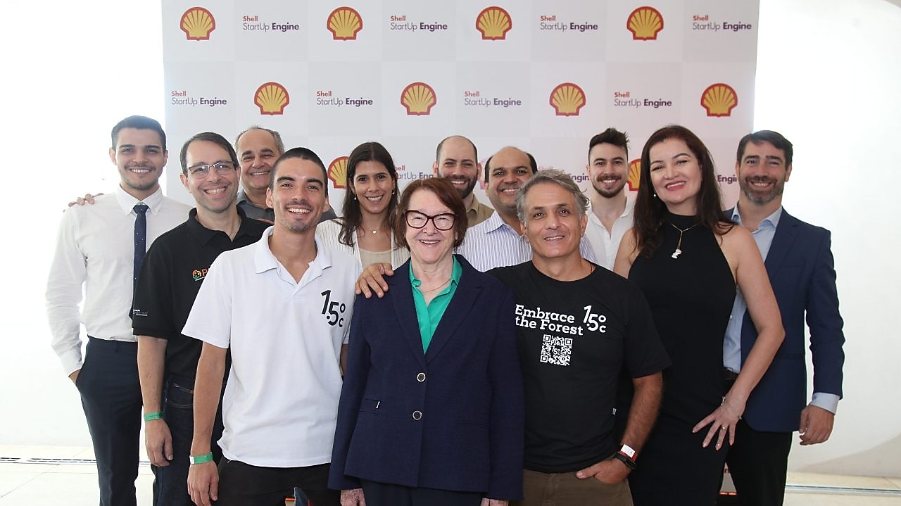 Grupo de 13 pessoas posando sorridentes em frente a um painel com o logo da Shell e a marca Shell StartUp Engine. A foto mostra diversidade de gênero e estilo, com pessoas vestindo roupas sociais. O ambiente é iluminado e transmite clima de celebração e colaboração