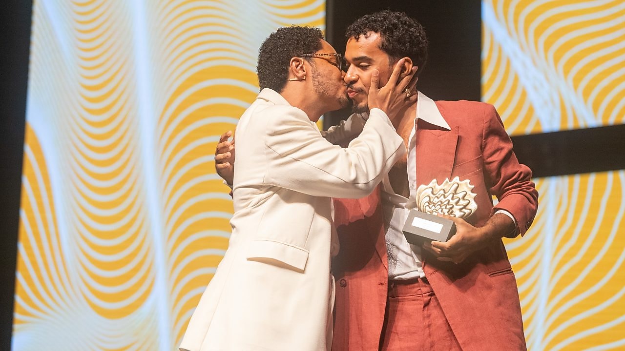 Dois homens se abraçam no palco durante uma premiação. O homem de terno branco e óculos dá um beijo carinhoso na bochecha do outro, que veste um terno vermelho e segura um troféu nas mãos. Ao fundo, um painel com padrão ondulado em amarelo e branco.