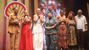 Grupo de pessoas no palco, algumas segurando microfones e um troféu, enquanto uma delas fala. Vestem roupas variadas, incluindo vestidos e conjuntos com estampas e cores vibrantes. Ao fundo, há o logotipo da Shell e uma projeção ampliada do grupo.
