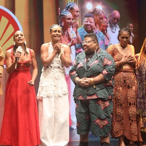 Grupo de pessoas no palco, algumas segurando microfones e um troféu, enquanto uma delas fala. Vestem roupas variadas, incluindo vestidos e conjuntos com estampas e cores vibrantes. Ao fundo, há o logotipo da Shell e uma projeção ampliada do grupo.