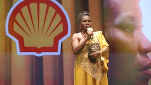 Pessoa no palco fala ao microfone enquanto segura um troféu em formato de concha. Usa um cocar com penas, veste roupa em tons de vermelho e preto e acessórios coloridos. Ao fundo, há iluminação quente e elementos dourados, sugerindo uma cerimônia ou premiação.