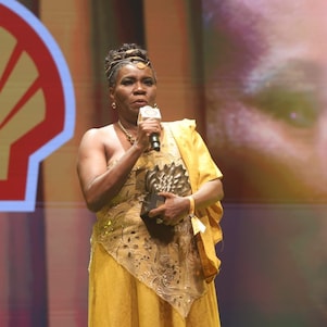 Pessoa no palco fala ao microfone enquanto segura um troféu em formato de concha. Usa um cocar com penas, veste roupa em tons de vermelho e preto e acessórios coloridos. Ao fundo, há iluminação quente e elementos dourados, sugerindo uma cerimônia ou premiação.