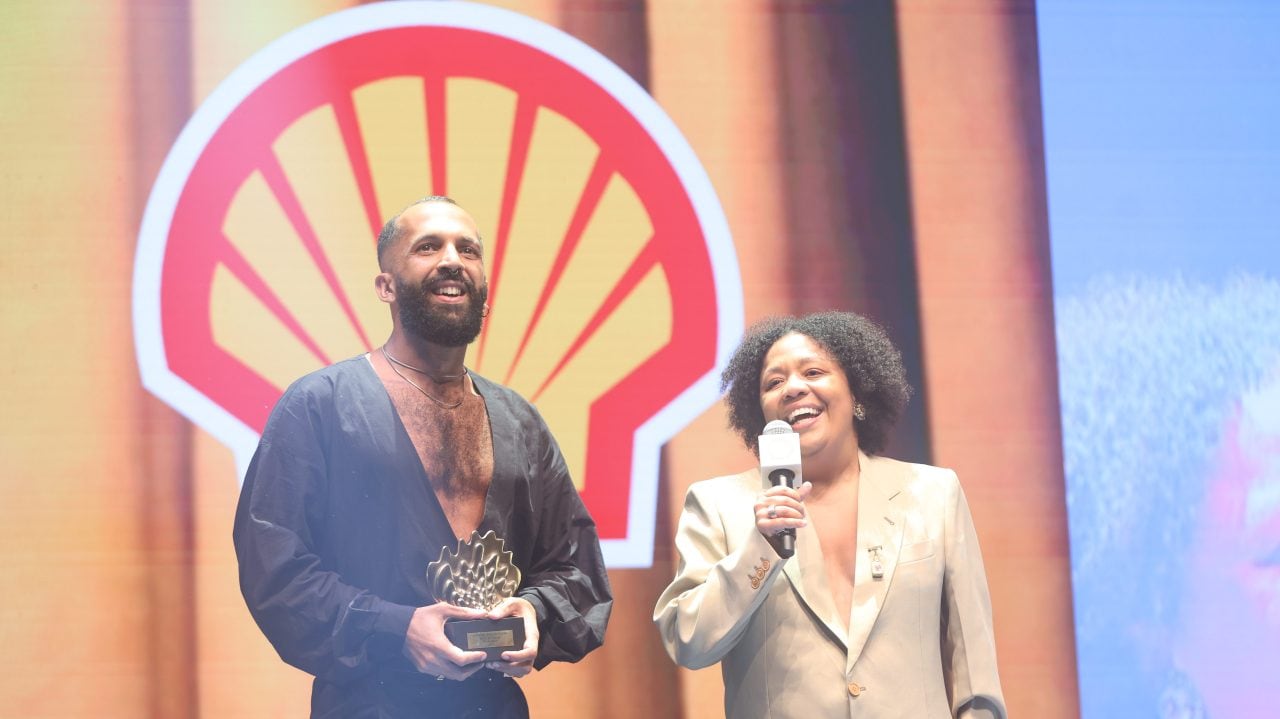 Duas pessoas em um palco: uma segura um troféu enquanto a outra fala ao microfone. Estão sorrindo e vestem roupas formais em tons escuros e claros. Ao fundo, há o logotipo da Shell e iluminação em tons quentes.