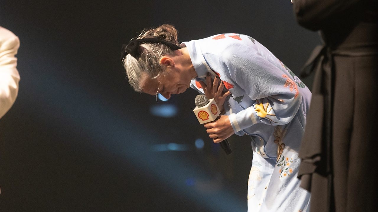 Uma mulher de cabelos brancos presos com uma faixa preta se inclina em um gesto de gratidão no palco do Prêmio Shell de Teatro. Ela segura um microfone com o logotipo da Shell em uma das mãos e coloca a outra sobre o peito, expressando emoção. Ela veste um vestido azul claro com bordados florais coloridos. A iluminação destaca sua presença no palco, enquanto outra pessoa parcialmente visível segura um troféu em formato de concha ao fundo. O momento transmite emoção e reconhecimento.