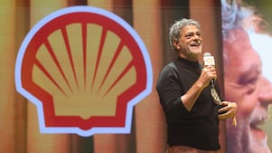 Pessoa em um palco segurando um microfone com a marca Shell e um troféu, sorrindo enquanto fala. Usa blusa preta de manga longa e calça em tom terroso. Ao fundo, há o logotipo da Shell e uma projeção ampliada de seu rosto.