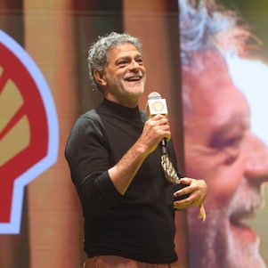 Pessoa em um palco segurando um microfone com a marca Shell e um troféu, sorrindo enquanto fala. Usa blusa preta de manga longa e calça em tom terroso. Ao fundo, há o logotipo da Shell e uma projeção ampliada de seu rosto.