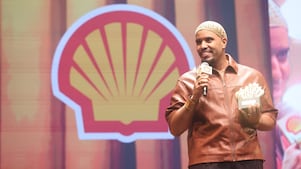 Pessoa em um palco segurando um microfone com a marca Shell e um troféu, enquanto fala. Usa uma jaqueta marrom e um gorro claro. Ao fundo, há o logotipo da Shell e uma projeção ampliada de seu rosto.