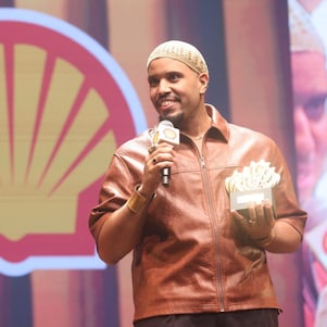 Pessoa em um palco segurando um microfone com a marca Shell e um troféu, enquanto fala. Usa uma jaqueta marrom e um gorro claro. Ao fundo, há o logotipo da Shell e uma projeção ampliada de seu rosto.