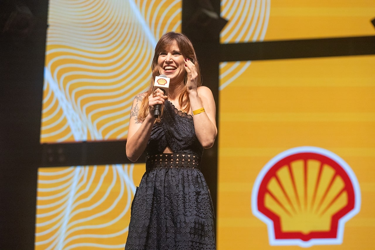 Mulher sorridente em palco segurando um microfone com o logotipo da Shell. Ela veste um vestido azul marinho rendado e está diante de um fundo iluminado em tons de amarelo com o símbolo da Shell em destaque. A cena parece ser de um evento corporativo ou premiação.