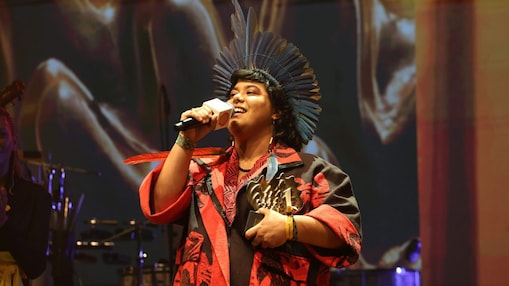 Pessoa no palco fala ao microfone enquanto segura um troféu em formato de concha. Usa um cocar com penas, veste roupa em tons de vermelho e preto e acessórios coloridos. Ao fundo, há iluminação quente e elementos dourados, sugerindo uma cerimônia ou premiação.