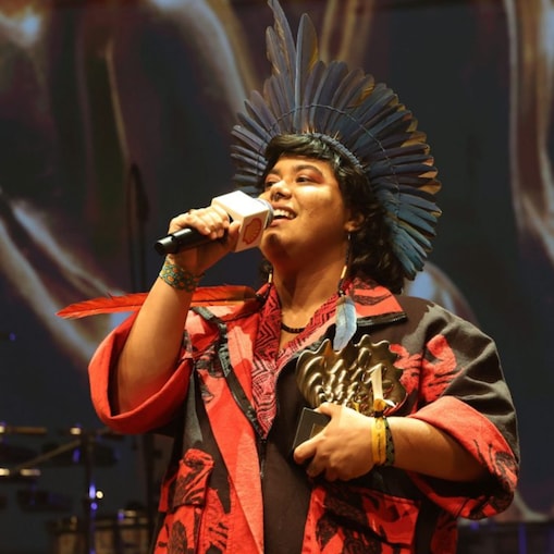 Pessoa no palco fala ao microfone enquanto segura um troféu em formato de concha. Usa um cocar com penas, veste roupa em tons de vermelho e preto e acessórios coloridos. Ao fundo, há iluminação quente e elementos dourados, sugerindo uma cerimônia ou premiação.