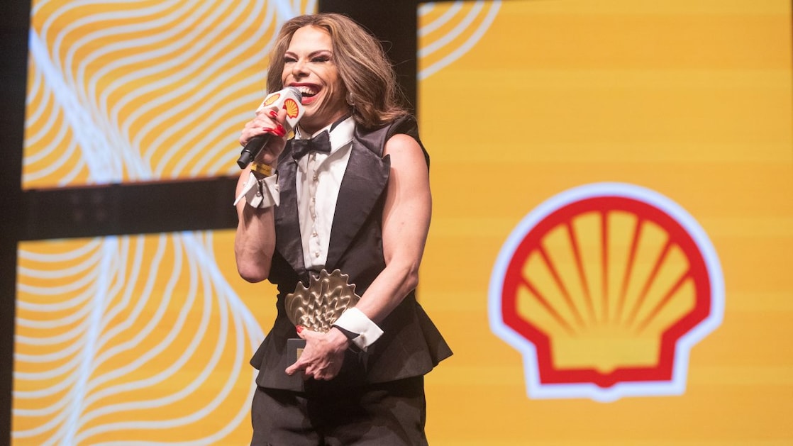 Drag queen sorridente segura um troféu em formato de concha dourada enquanto fala ao microfone no palco do Prêmio Shell de Teatro. Ela usa terno preto com camisa branca e gravata borboleta, em frente a um telão com o logotipo da Shell sobre fundo amarelo com listras brancas curvas.